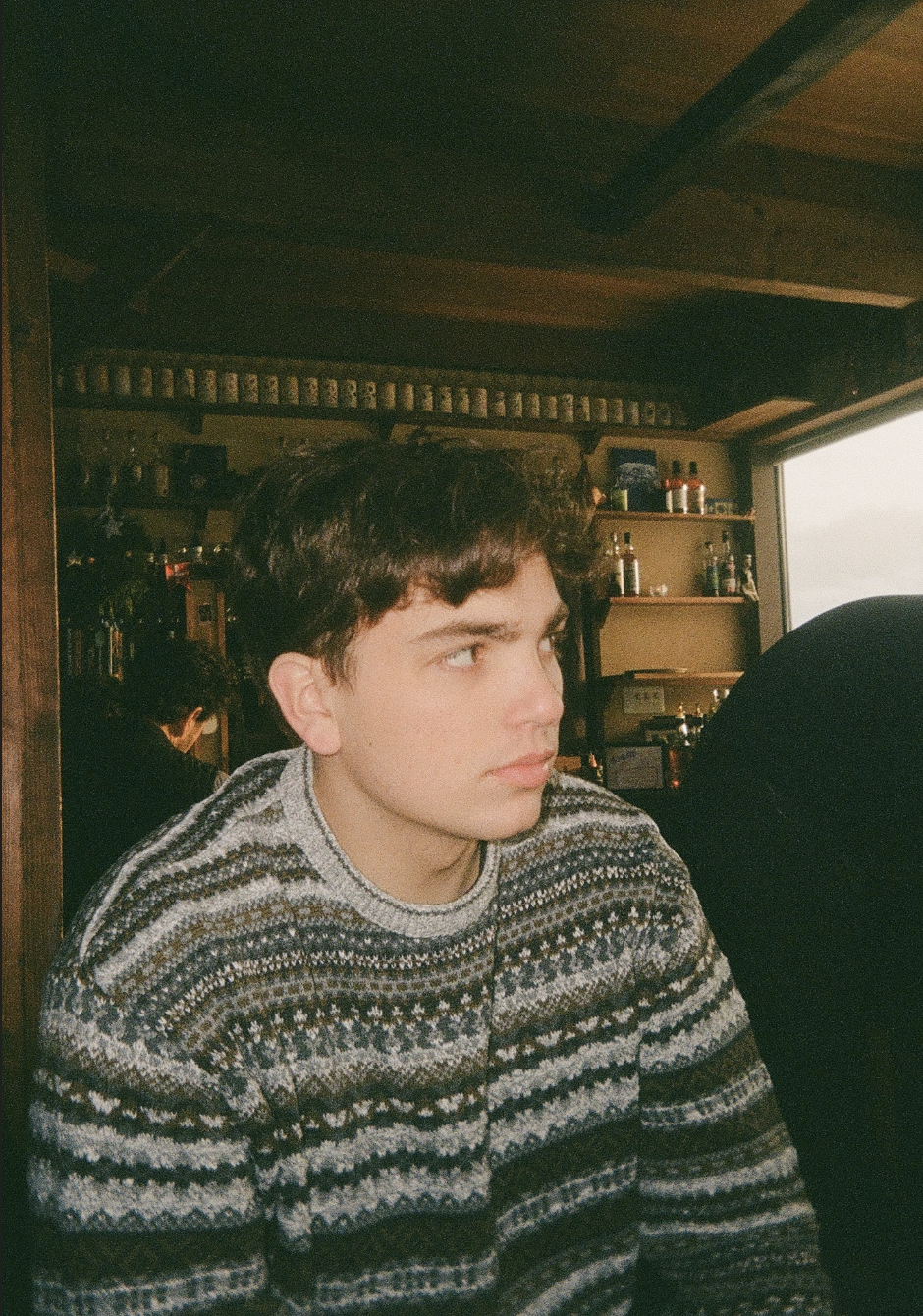 A young man, likely in his late teens or early twenties, with a distinctive, symmetrical face and strong features. He has dark, slightly messy hair styled casually, giving him a contemporary yet timeless look. His expression is pensive and slightly melancholic, with a distant, thoughtful gaze. He has strong eyebrows, a straight nose, and defined cheekbones. The lighting from a window casts soft shadows that highlight his features. His overall appearance aligns with current 'Gen Z' trends.